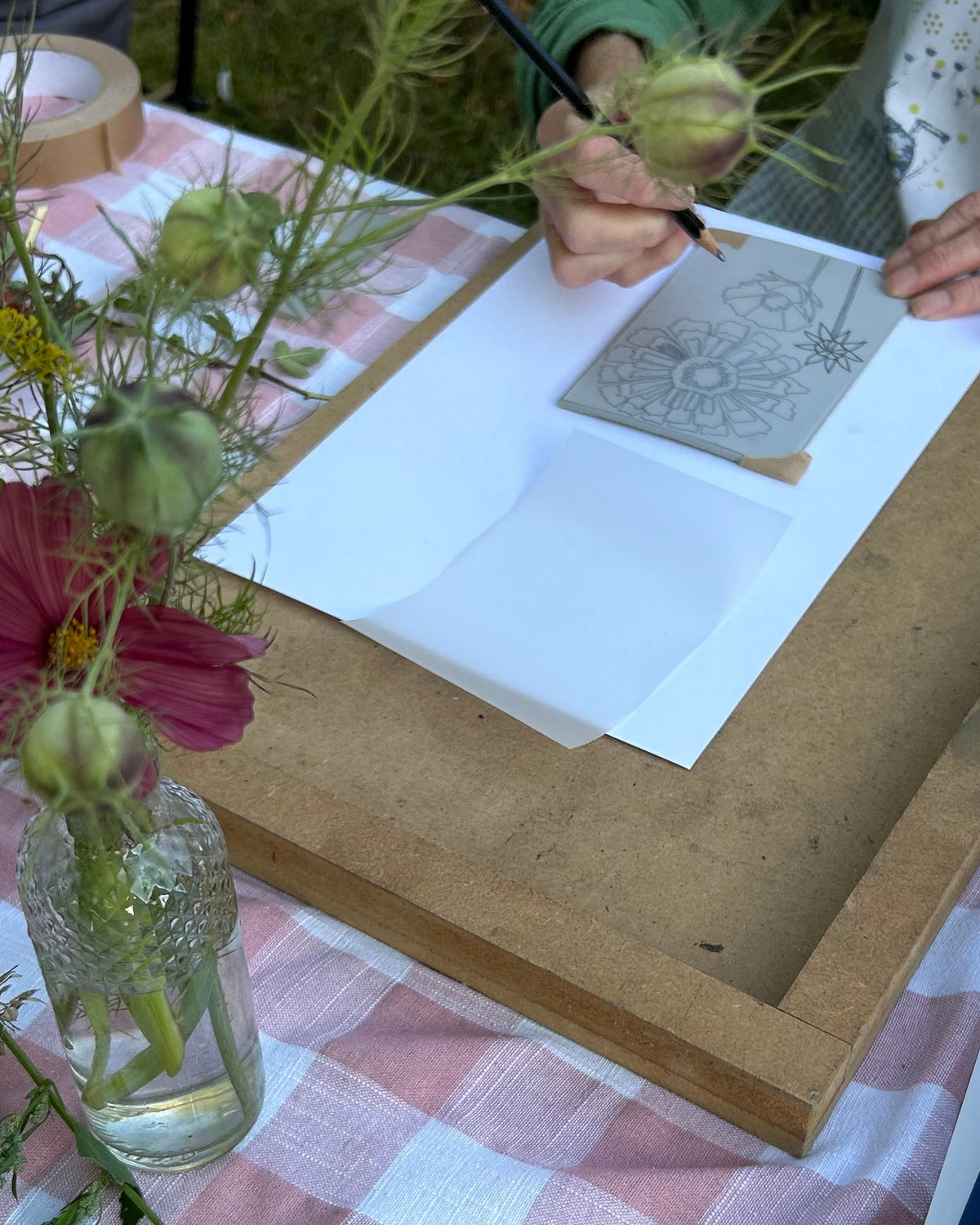 Person drawing on paper at a table with flowers and a checkered tablecloth.