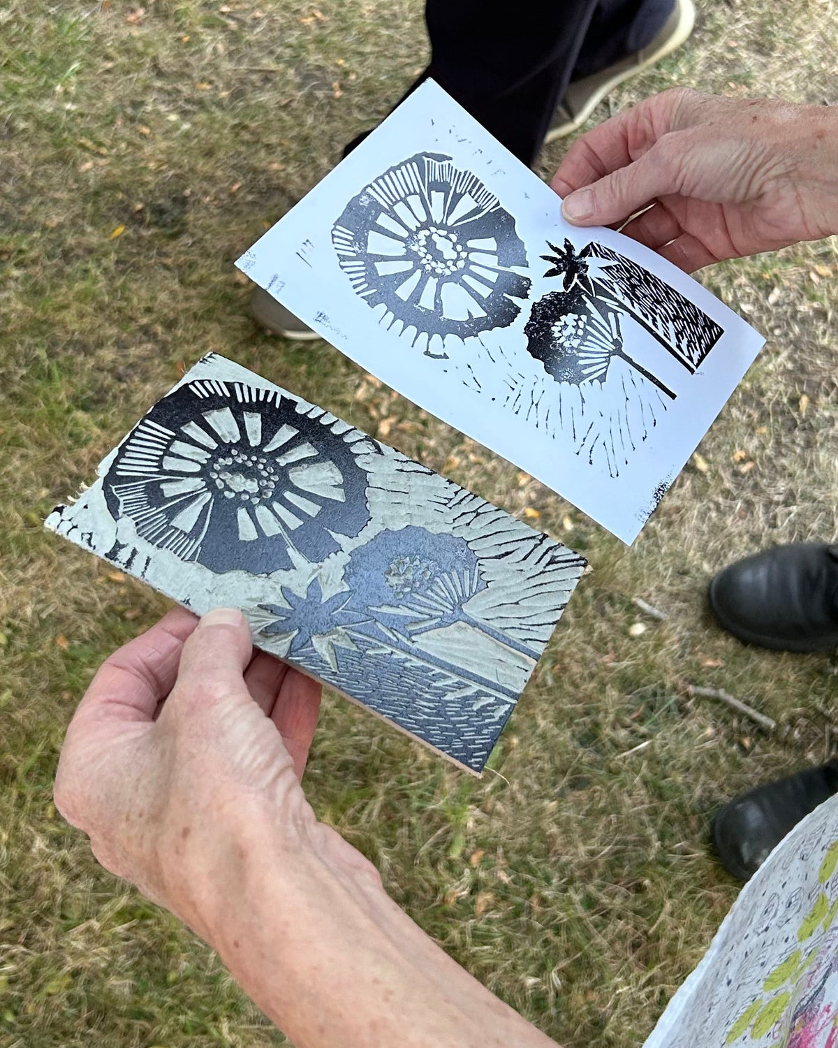 Two people holding stencils with floral designs outdoors on a grassy area.