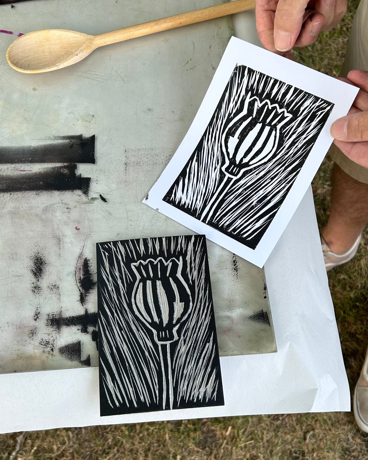 Person holding a print of a black and white tulip design on a white sheet, with another print and a wooden spoon in the background.