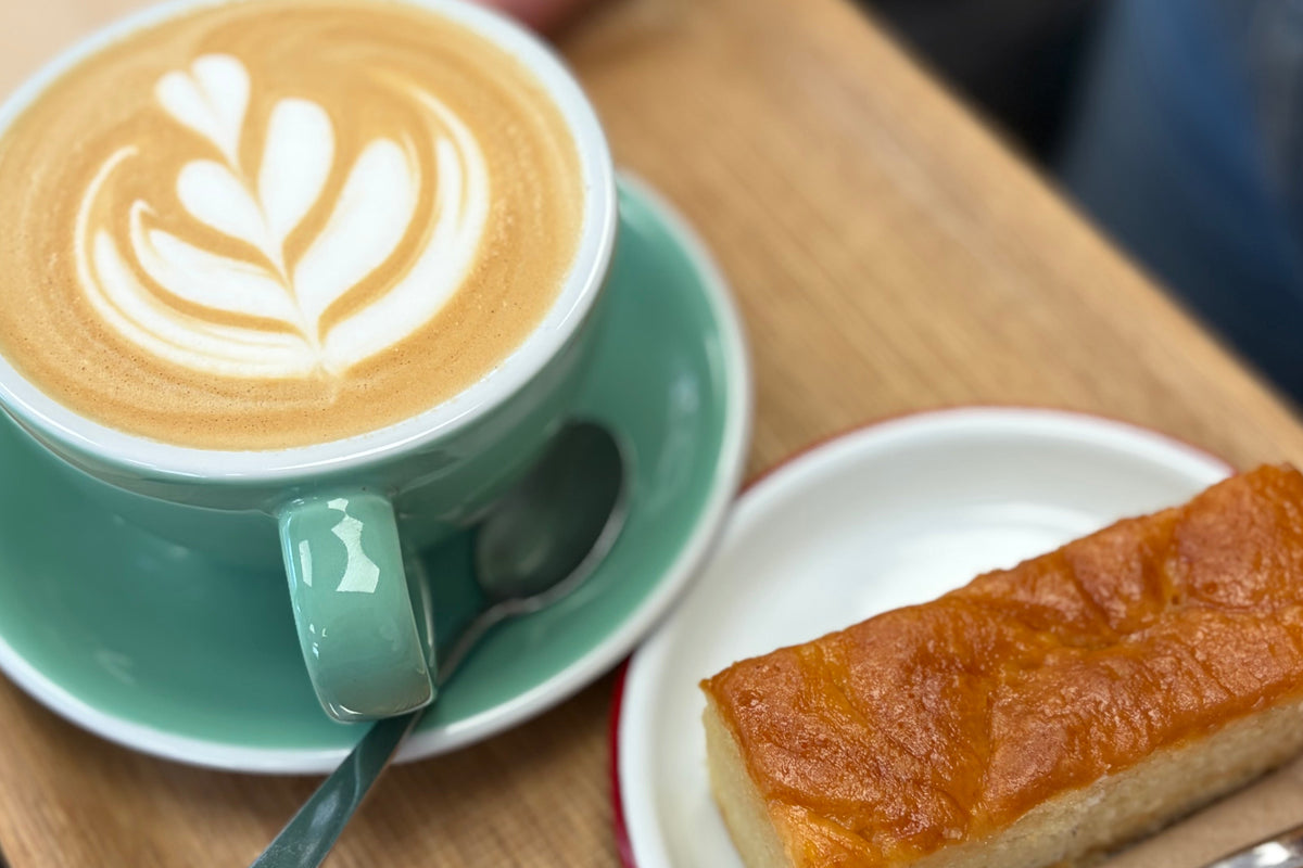 A wooden tray held by a person in a cafe setting, featuring a cup of latte with leaf-shaped latte art on a mint green saucer, and a slice of golden-brown cake on a small white plate with a fork and napkin.