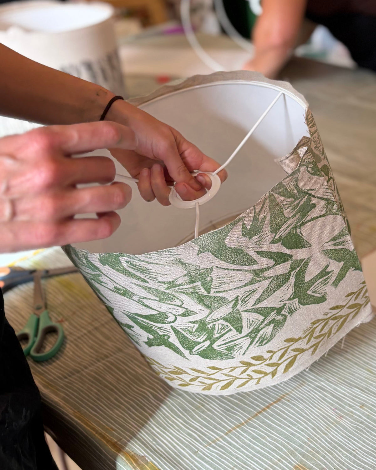 A person's hands are shown wrapping white cord around a paper lampshade with a green botanical pattern and a gold patterned band. The scene is set on a rustic wooden table with scissors, paper, and other crafting materials, with other people working in the background.