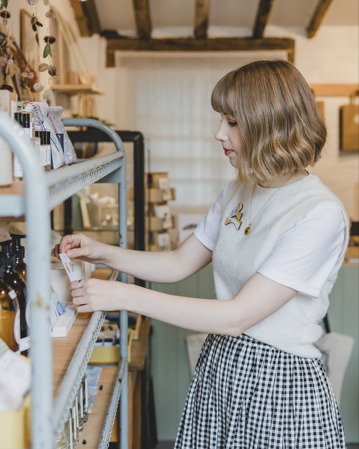 A woman with short blonde hair stands in a cozy, rustic shop or studio, wearing a white t-shirt and a black-and-white gingham skirt. She is reaching for items on a metal shelving unit filled with bottles and packaged goods.