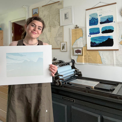 Person holding a print in front of a large printing press with artwork on the walls.